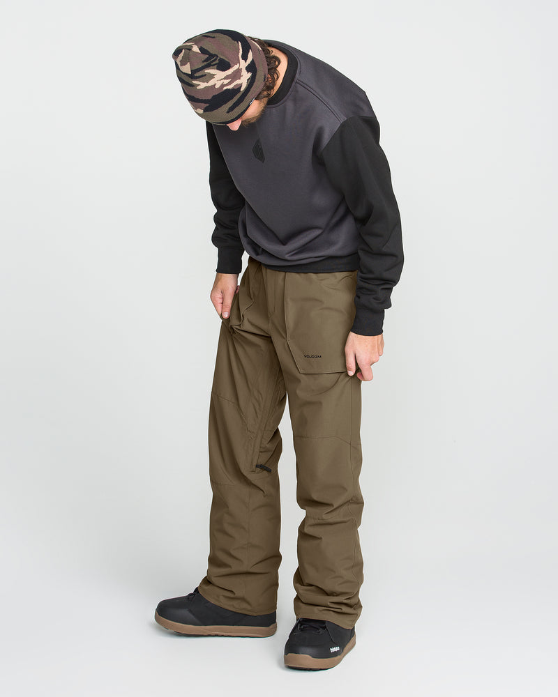 A person dressed in a camo beanie, dark sweatshirt, and Volcom Men’s Roan 20K Pants—crafted from recycled fabric in olive green—stands looking down against a plain light background.