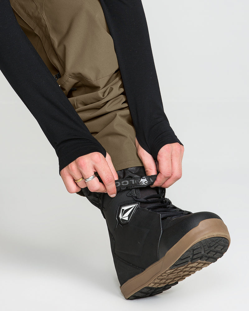 A person wearing Volcom Men’s Roan 20K Pants in brown, made from recycled fabric, and a black long-sleeve top fastens the strap on a black snowboarding boot with a brown sole and a geometric logo.