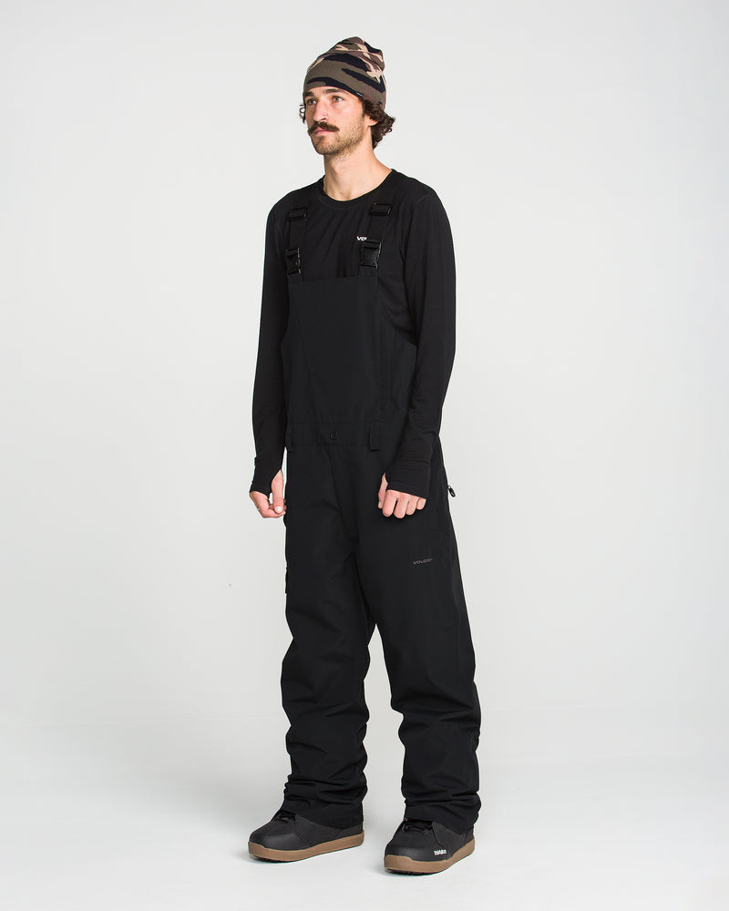 A person wearing a black beanie, a black long-sleeve shirt, and the Volcom Men’s V.Co Sparta Bib—crafted from recycled fabric with waterproof protection—stands against a plain white background.