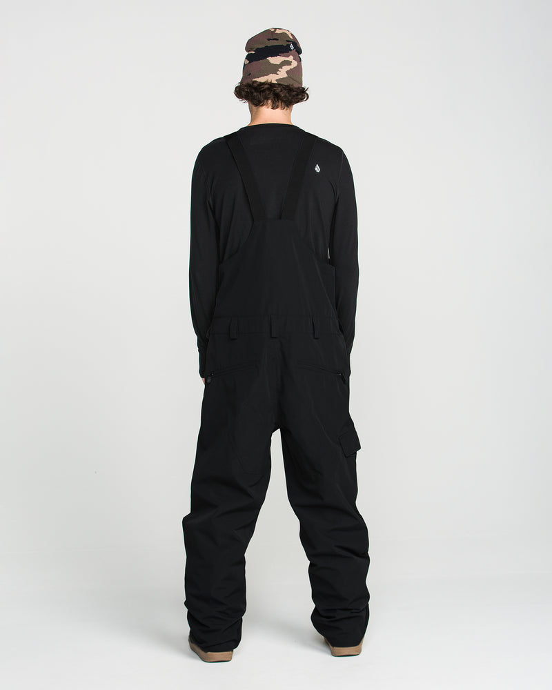 A person stands facing away, wearing the Volcom Men’s V.Co Sparta Bib in black—crafted from recycled fabric—paired with a black long-sleeve shirt and a camouflage bucket hat. The background is plain white.
