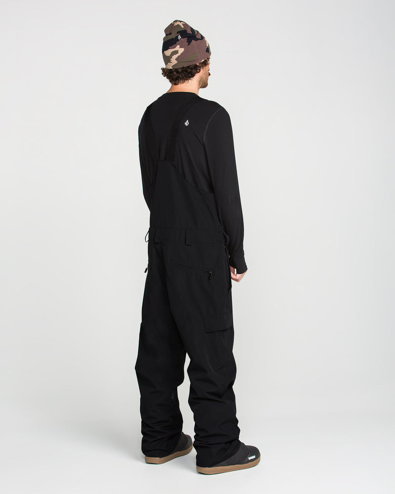 Wearing the Volcom Men’s V.Co Sparta Bib crafted from recycled fabric, a person stands facing away against a plain white background, paired with a black long-sleeve shirt, black boots, and a camouflage beanie.