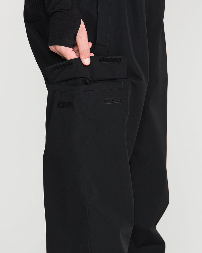 Wearing Volcom’s Men’s V.Co Sparta Bib in black—crafted from recycled fabric—a person opens a rectangular side pocket, fastened with velcro strips, revealing their hand partially inside the pocket. Only the lower torso and hand are visible.