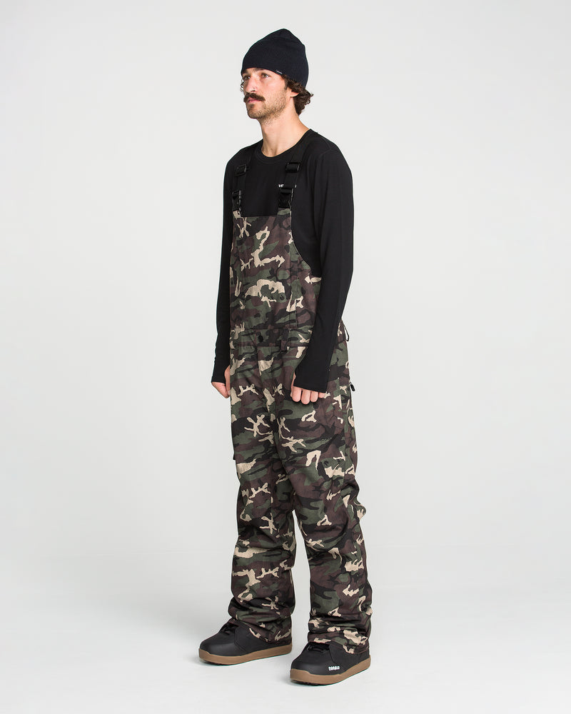 A person wearing a black beanie, long-sleeve black shirt, and the Volcom Men’s V.Co Sparta Bib—baggy camo overalls made from recycled fabric—stands against a plain white background, looking slightly to the side.