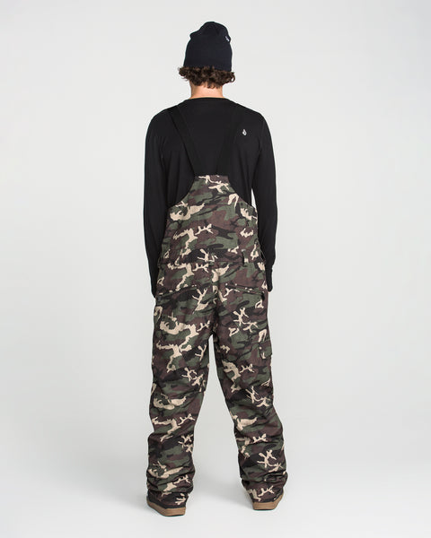 A person stands with their back to the camera on a plain white background, wearing a black long-sleeve shirt, brown shoes, a black beanie, and the Volcom Men’s V.Co Sparta Bib camo overalls made from recycled fabric.