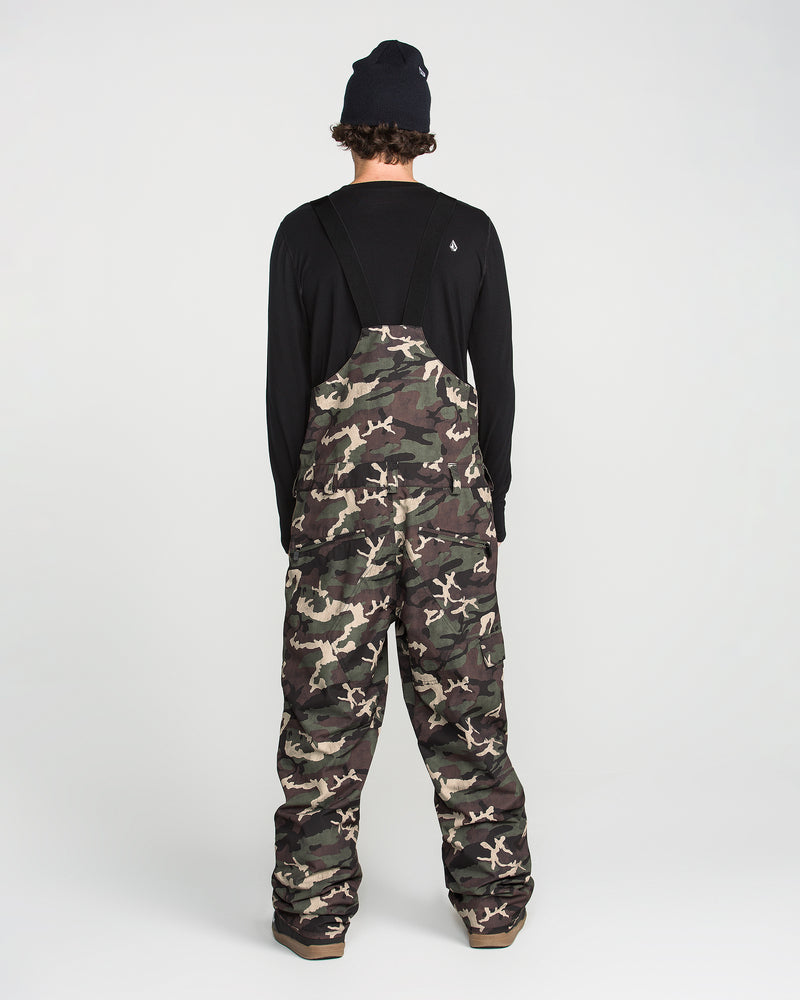 A person stands with their back to the camera on a plain white background, wearing a black long-sleeve shirt, brown shoes, a black beanie, and the Volcom Men’s V.Co Sparta Bib camo overalls made from recycled fabric.