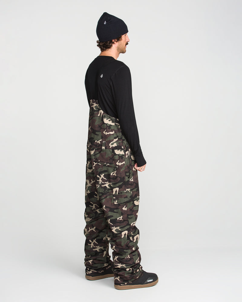 A person stands sideways on a white background wearing the Volcom Men’s V.Co Sparta Bib—camouflage, baggy overalls made from recycled fabric—paired with a black long-sleeve shirt and black beanie.