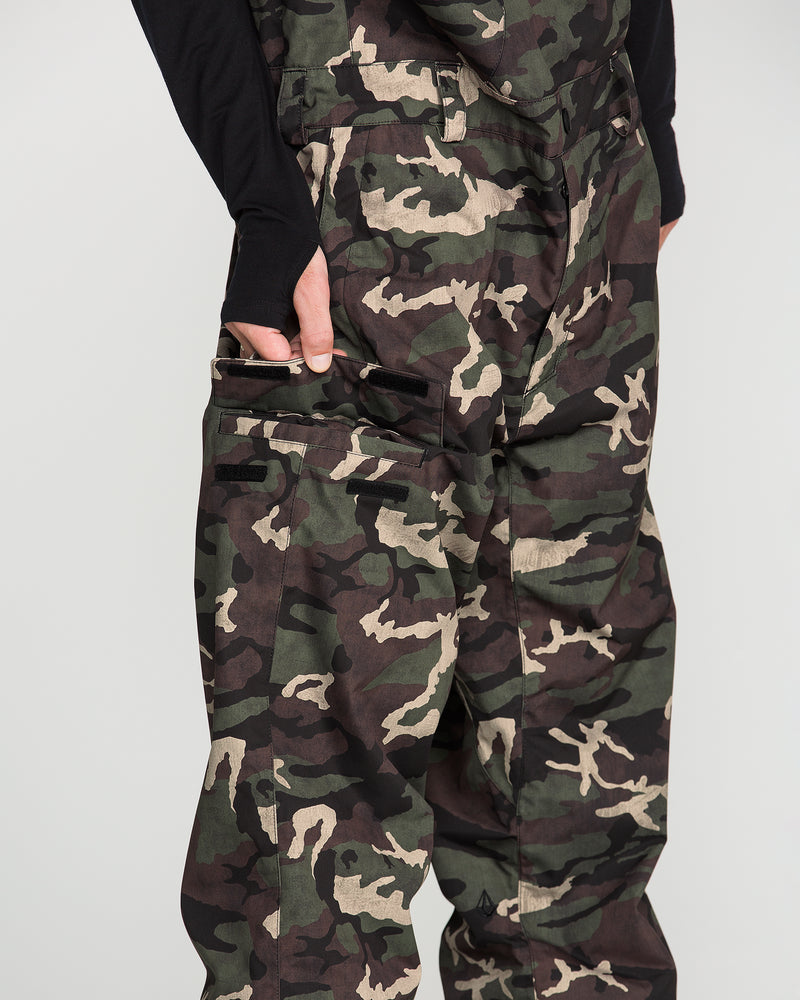 A person wearing the Volcom Men’s V.Co Sparta Bib in camouflage, made from recycled fabric, paired with a black long-sleeve shirt and standing with their left hand in the bib pocket against a plain light background.
