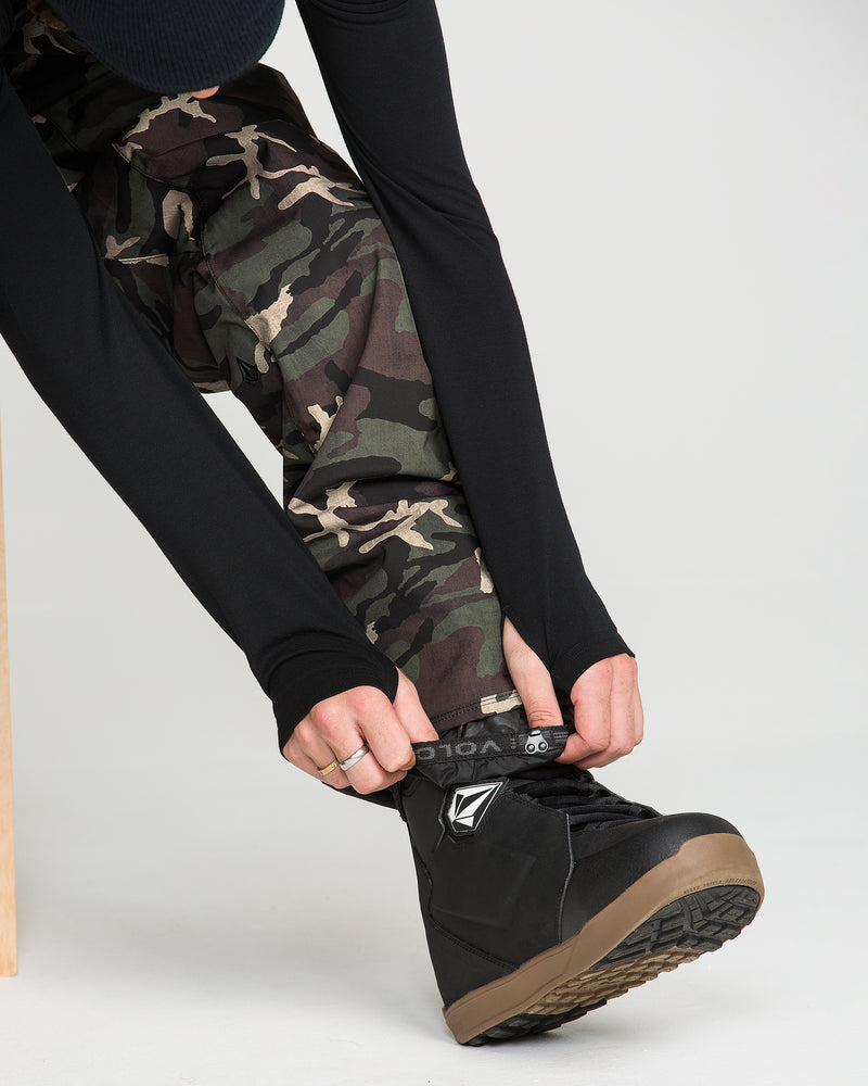 A person wearing the Volcom Men’s V.Co Sparta Bib in camouflage print and a black long-sleeve top sits and tightens the strap on a black snowboarding boot.