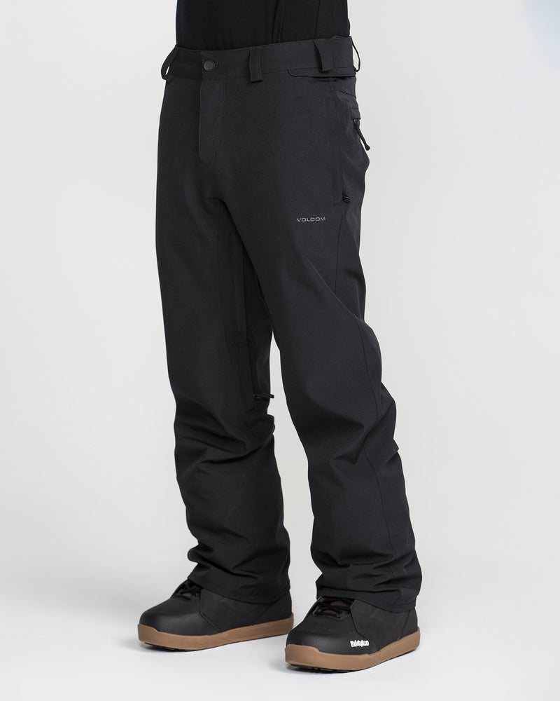 A person wearing black Volcom Men’s Freakin Snow Chino Pants and black winter boots with brown soles stands against a plain light background. The photo focuses on the lower half of the body, accentuating the men’s ski pants crafted from waterproof, breathable fabric by Volcom.