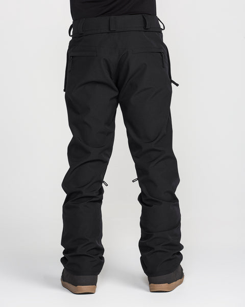 A person stands facing away, showcasing the Volcom Men’s Freakin Snow Chino Pants in black with brown boots against a plain white background. Only the lower body is visible, emphasizing the waterproof and breathable fabric of these men’s ski pants.