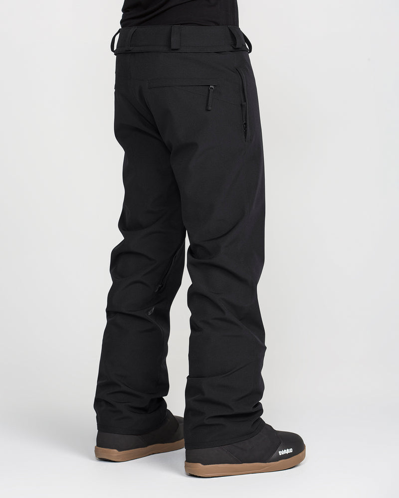 A person wearing Volcom Men’s Freakin Snow Chino Pants in black and black winter boots with tan soles stands on a white background, viewed from behind. These waterproof, breathable pants from Volcom provide comfort and protection in cold weather.