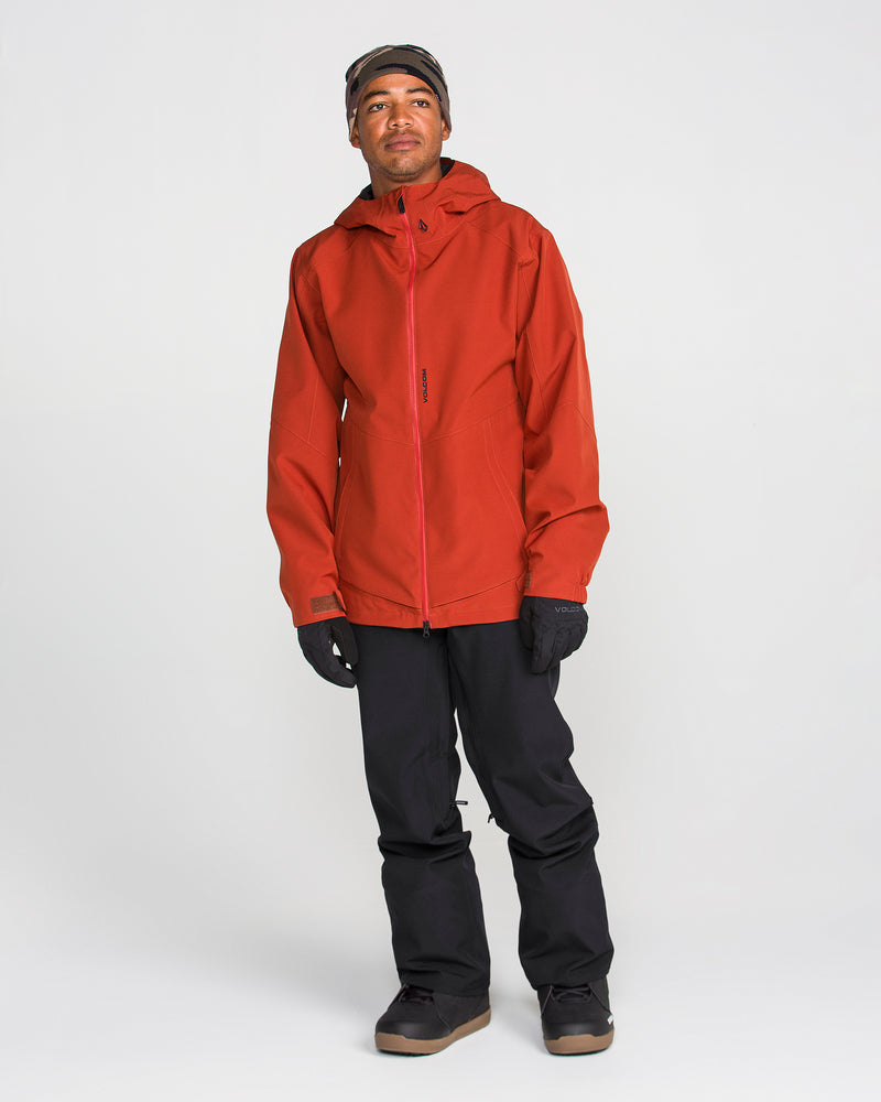 A person wearing a red hooded jacket, gloves, and winter boots stands against a plain white background, looking slightly upward. They are wearing Volcom Men’s Freakin Snow Chino Pants, whose waterproof breathable fabric ensures comfort and protection in wintry conditions.