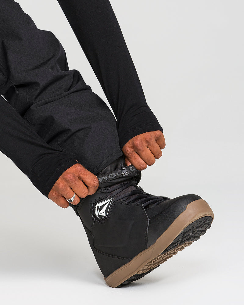 A person wearing Volcom Men’s Freakin Snow Chino Pants in black adjusts the cuff over a black snowboarding boot with a brown sole on a light background.