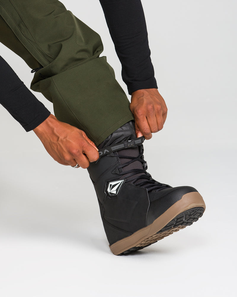 A person wearing Volcom Men’s Freakin Snow Chino Pants in green and a black shirt tightens the laces on a black snowboarding boot with a brown sole against a plain light background.