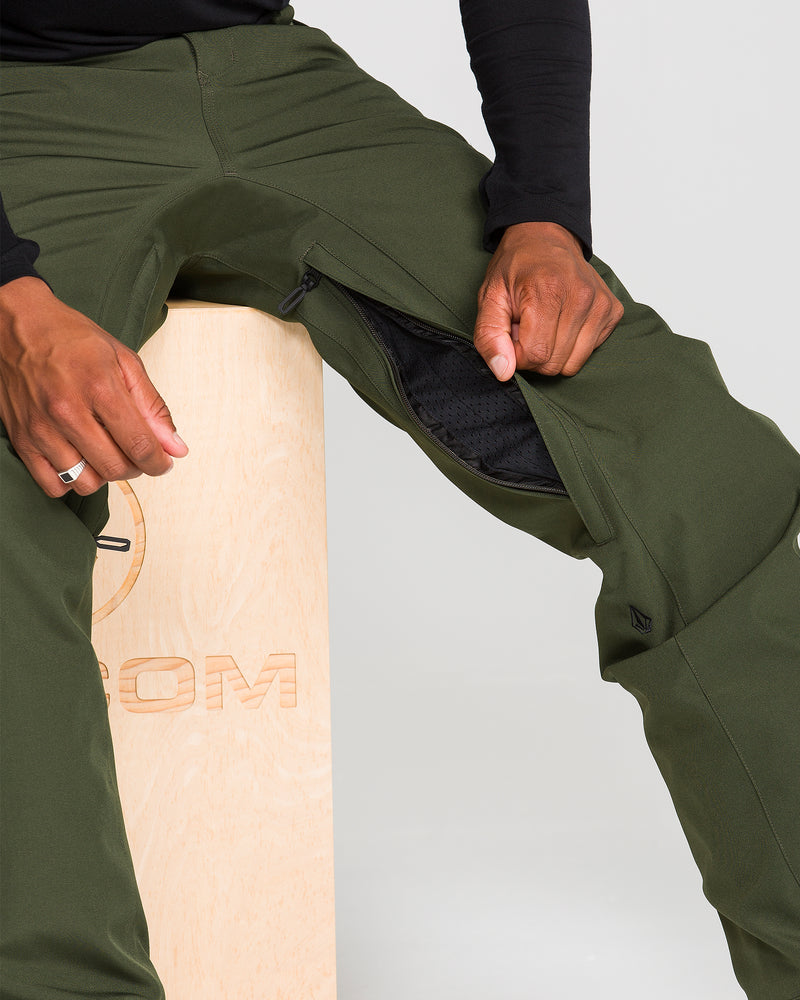 A person sits on a wooden box, wearing Volcom Men’s Freakin Snow Chino Pants in green with the zippered thigh vent partially open to reveal black mesh lining inside. They pair the pants with a black long-sleeve shirt and a silver ring.