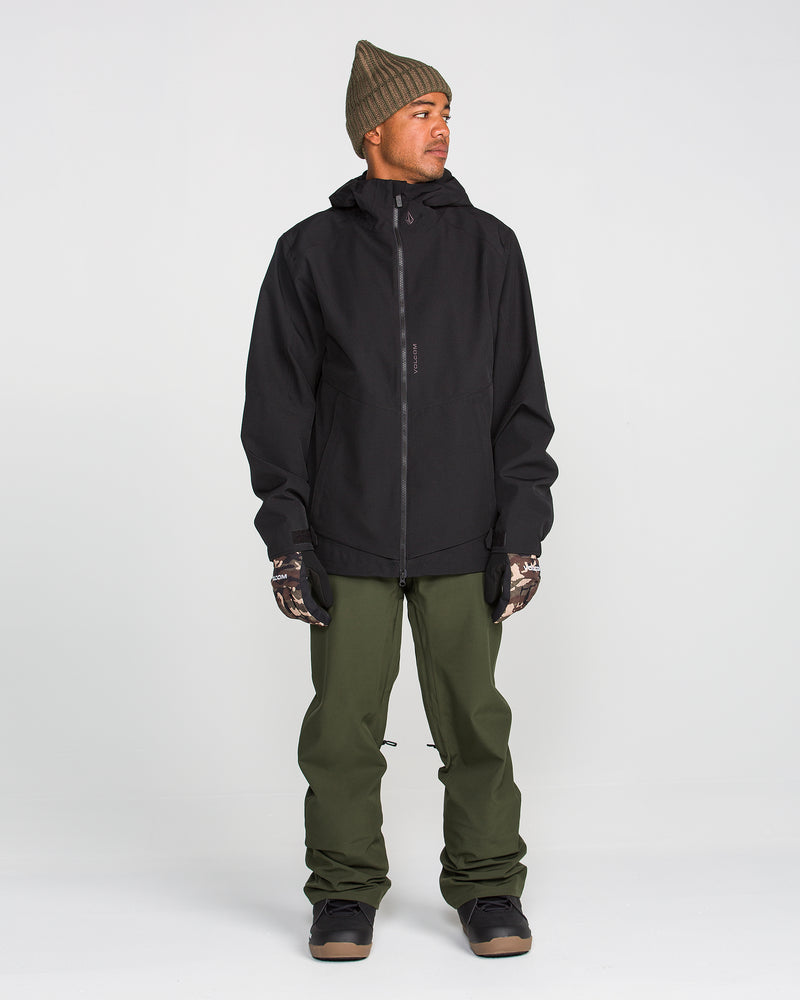 A person stands against a plain background, wearing a black hooded jacket, Volcom Men’s Freakin Snow Chino Pants in olive green, camouflage gloves, dark boots, and a brown knit beanie. The waterproof and breathable Volcom pants complete the look as the person gazes to the side with hands by their sides.