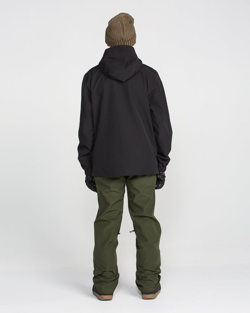 A person stands facing away, wearing a brown beanie, black hooded jacket, Volcom Men’s Freakin Snow Chino Pants in waterproof, breathable green, and gloves against a plain white background.