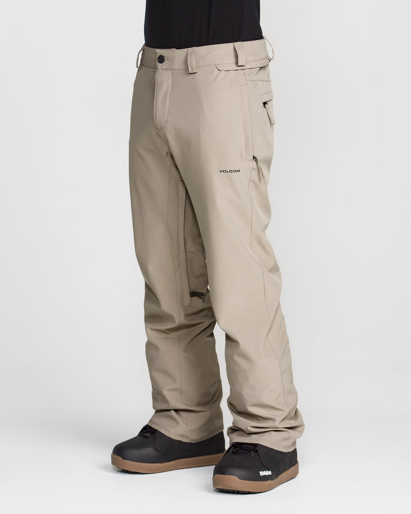 A person is shown from the waist down, wearing Volcom Men’s Freakin Snow Chino Pants in beige—crafted from recycled stretch Oxford fabric—and black snow boots, standing against a plain white background. The waterproof pants have a relaxed fit and zippered pockets.