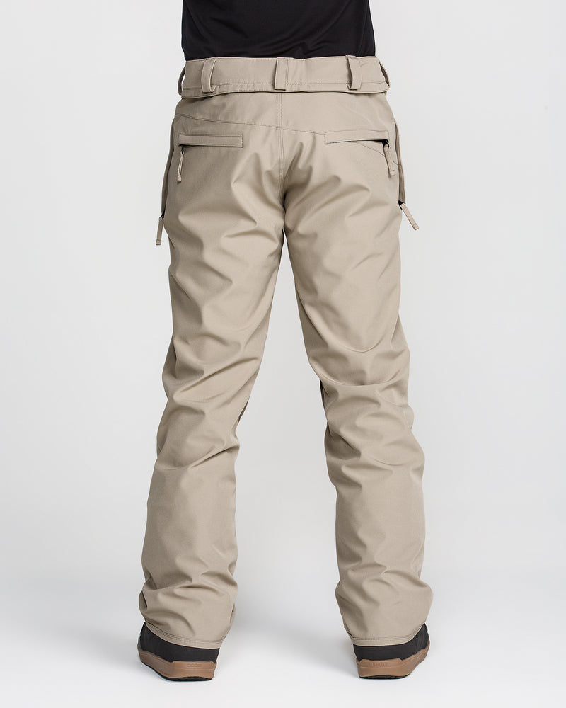 A person is shown from the back against a plain white background, wearing Volcom Men’s Freakin Snow Chino Pants in beige paired with brown boots. Only the lower half of the body is visible. The pants, made from recycled stretch Oxford material, include zipper pockets and belt loops.