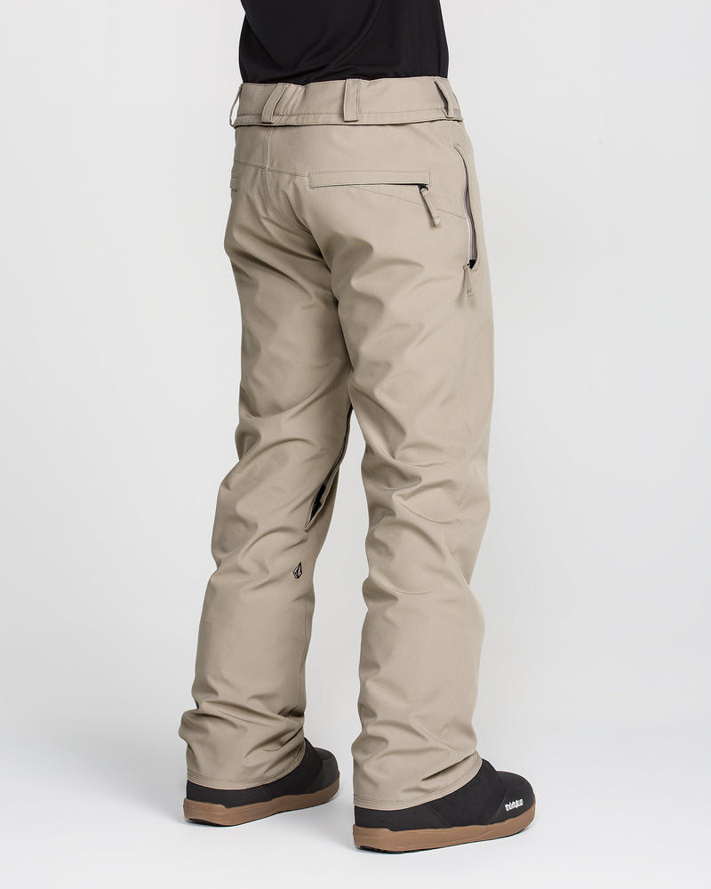 A person wearing Volcom Men’s Freakin Snow Chino Pants in beige and black boots stands facing away, revealing the back view with pockets and seam detail. These pants feature a waterproof, breathable design ideal for outdoor activities. The background is plain white.