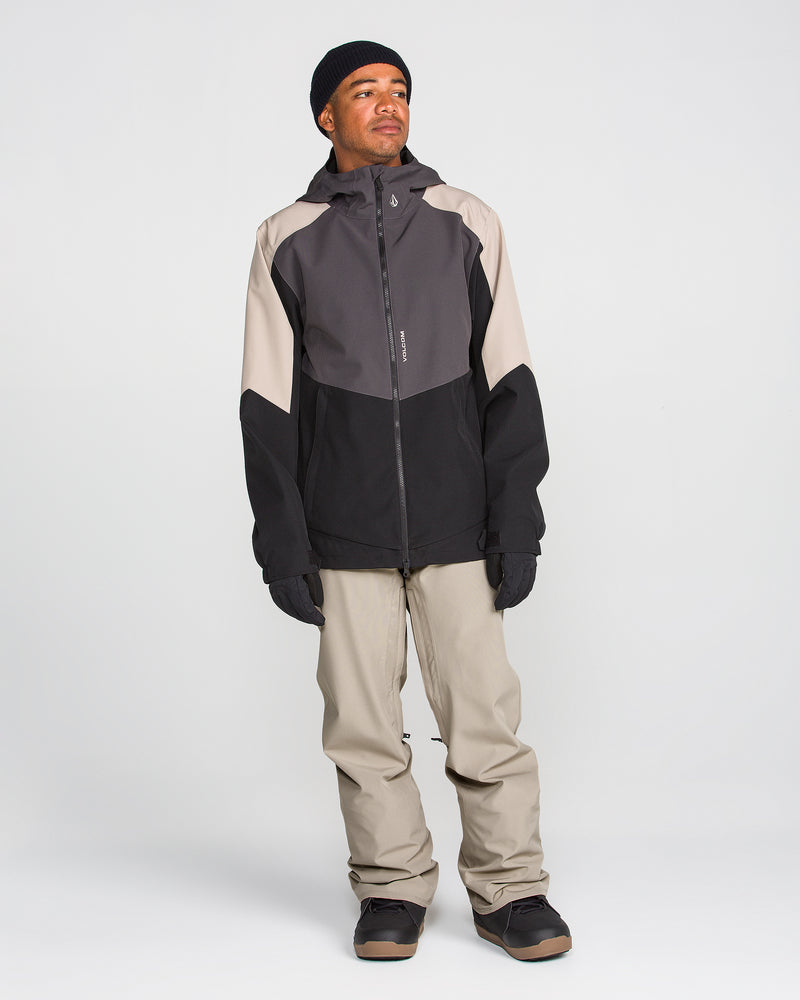 A person stands facing slightly left, wearing a black and gray hooded jacket, Volcom Men’s Freakin Snow Chino Pants in light beige that are waterproof and breathable, black gloves, a black beanie, and dark shoes against a plain white background.