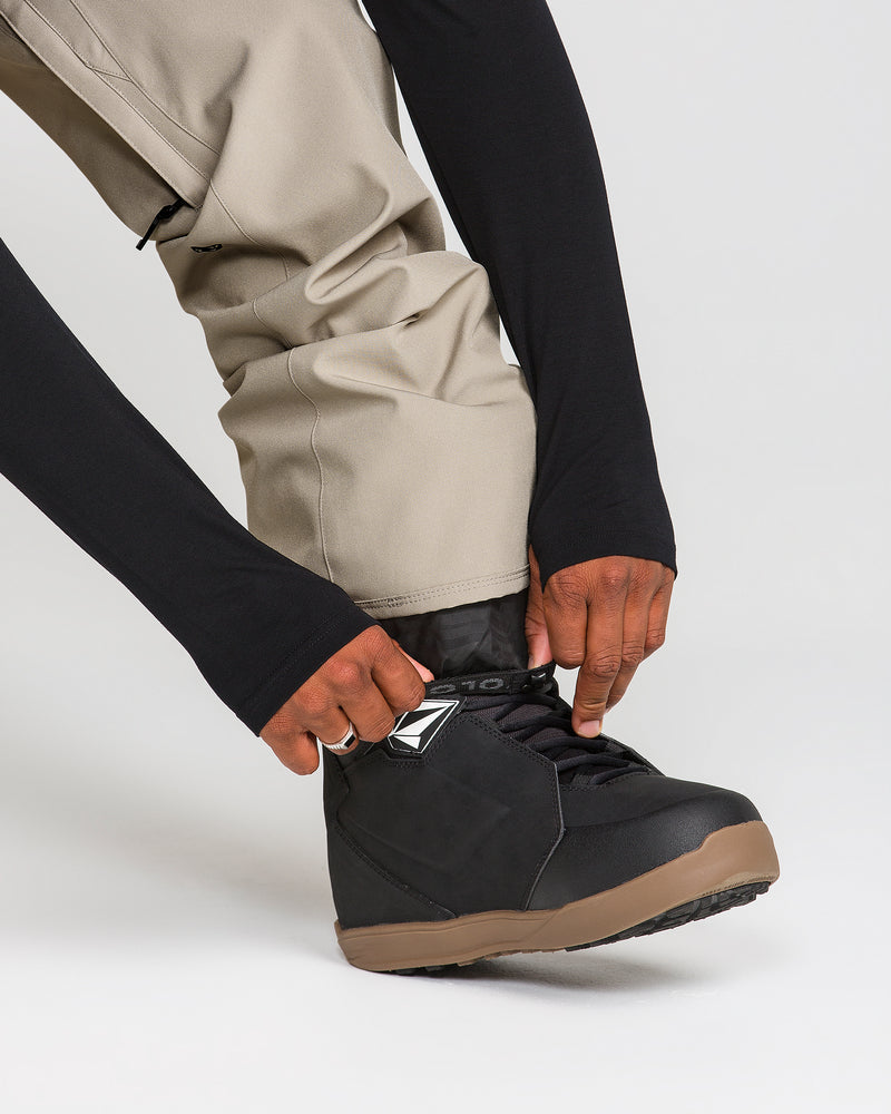 A person wearing Volcom Men’s Freakin Snow Chino Pants in beige and a black long-sleeve shirt is bending down to tie the laces of a black and brown shoe or boot.