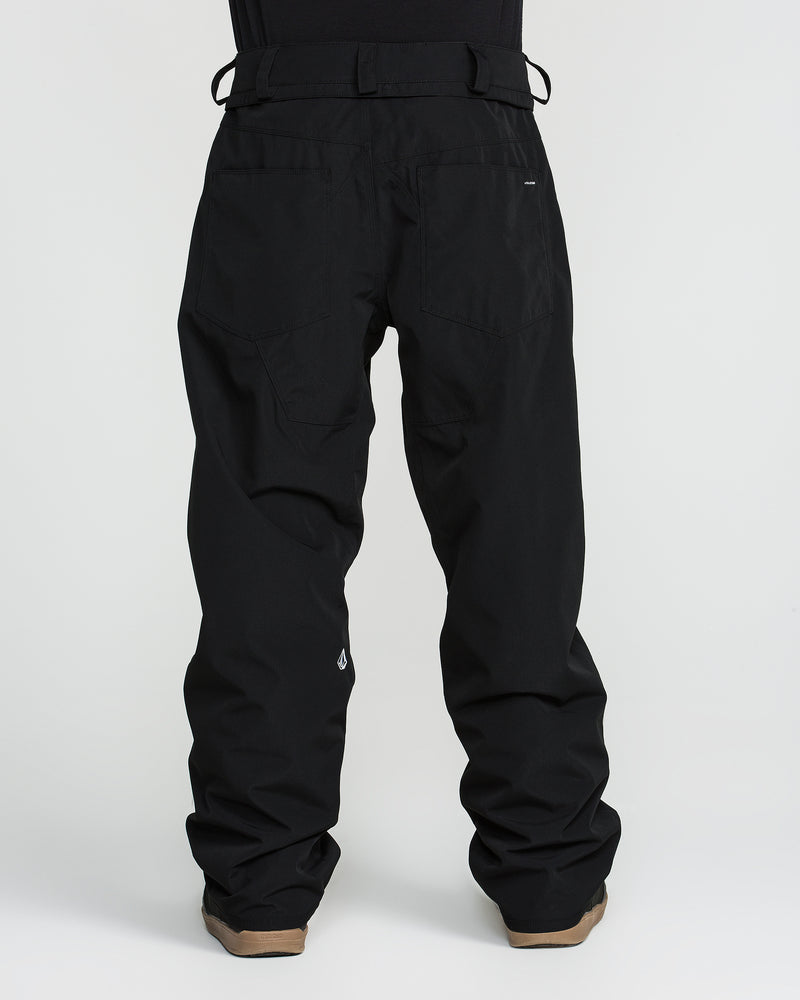 A person is shown from the back against a plain white background wearing Volcom Men’s Nailer Pants—loose-fitting black waterproof pants made from recycled materials—and brown shoes.