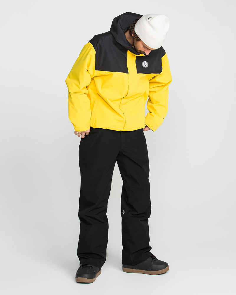 A person wearing a yellow and black jacket, black Volcom Men’s Nailer Pants made from recycled materials, black shoes, and a white beanie stands looking down against a plain white background.