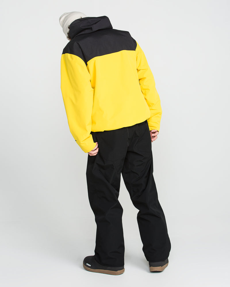 Wearing Volcom Men’s Nailer Pants—black waterproof pants made from recycled materials—a person in a yellow and black jacket, gray shoes, and a light-colored beanie stands facing away from the camera against a plain white background.