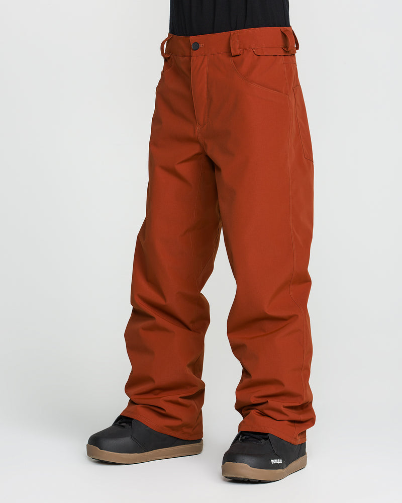 Wearing Volcom Men’s Nailer Pants in rust, crafted from recycled fabric, and black snow boots, the individual stands against a plain white background with only their lower body visible.