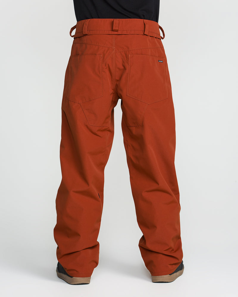 A person is shown from the back wearing rust-colored Volcom Men’s Nailer Pants against a plain white background. Crafted from recycled fabric, these pants feature belt loops and two back pockets, styled here with brown shoes for a classic look.