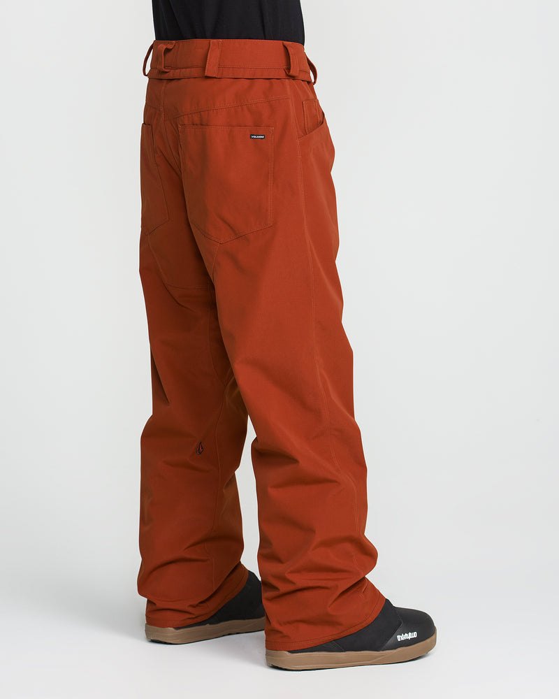 A person is shown in profile, displaying only the lower half of their body, wearing burnt orange Volcom Men’s Nailer Pants made from recycled fabric and paired with black boots against a white background.
