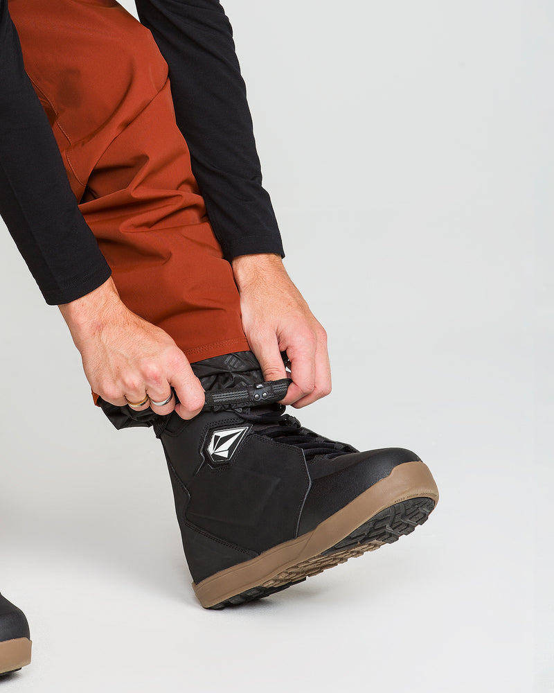 A person wearing rust-colored Volcom Men’s Nailer Pants, made from recycled fabric and designed to be waterproof, adjusts the top of a black snowboarding boot with a tan sole against a plain light background.