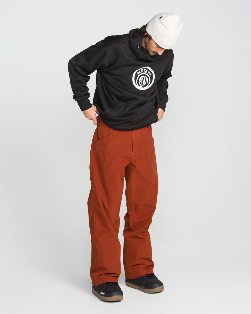 A person looks down while standing against a plain white background, wearing a white beanie, a black hooded sweatshirt featuring a circular logo, and loose-fitting red Volcom Men’s Nailer Pants made from recycled fabric.