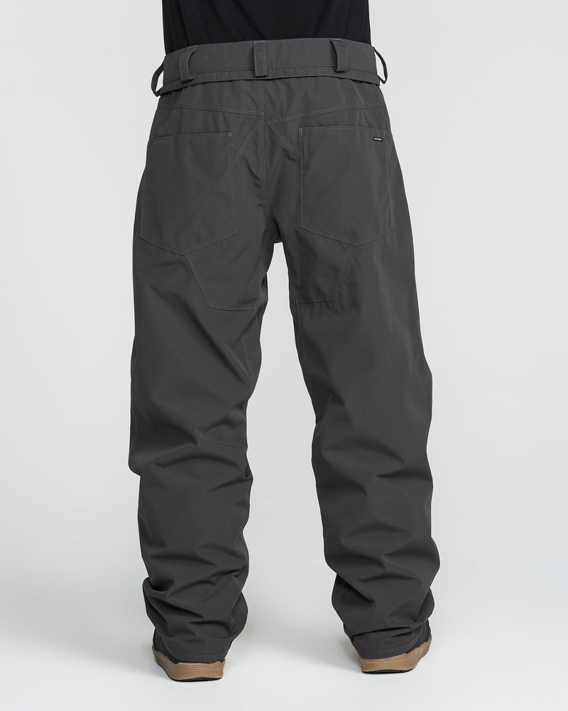 A person is shown from the back against a plain white background wearing Volcom Men’s Nailer Pants in dark gray, which are loose-fitting, waterproof, and crafted from recycled fabric. The pants feature large back pockets, sturdy belt loops, and are paired with tan shoes.