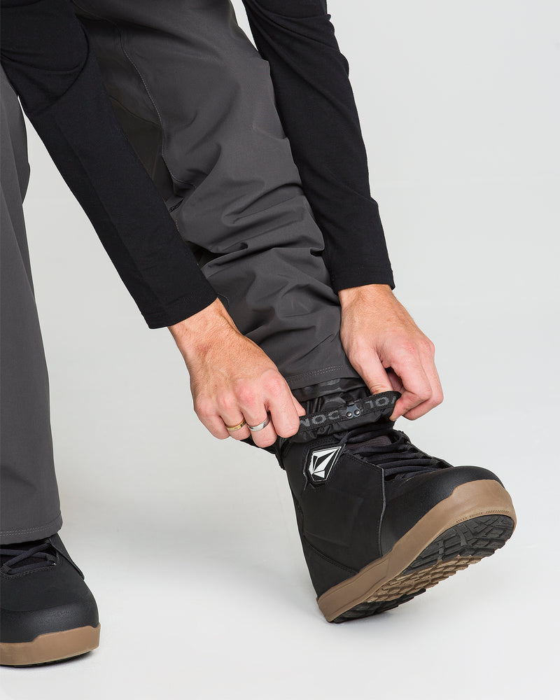 A person adjusts the strap on a black snow boot over Volcom Men’s Nailer Pants in gray, preparing for outdoor activity. The photo focuses on the hands, the boot, and part of the lower legs against a plain background.