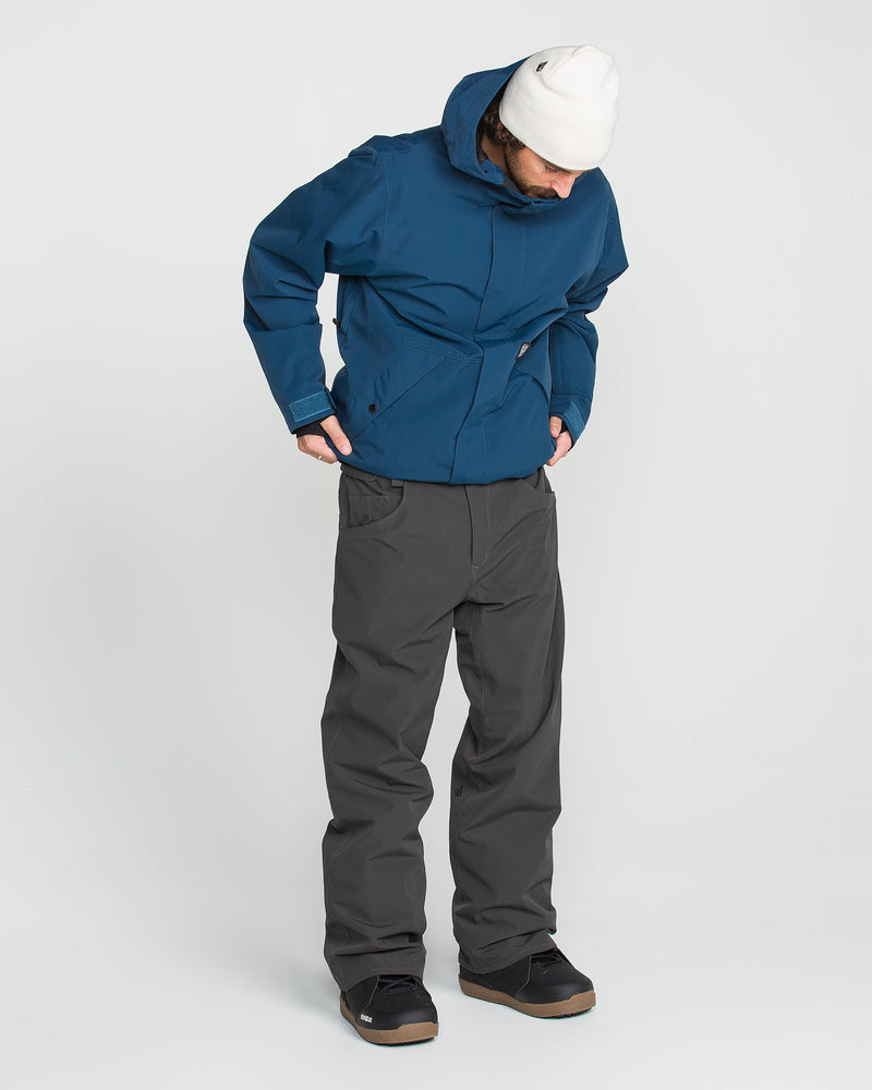 A person wearing a blue hooded jacket, Volcom Men’s Nailer Pants, black shoes, and a white beanie looks down with hands in their pockets against a plain white background.