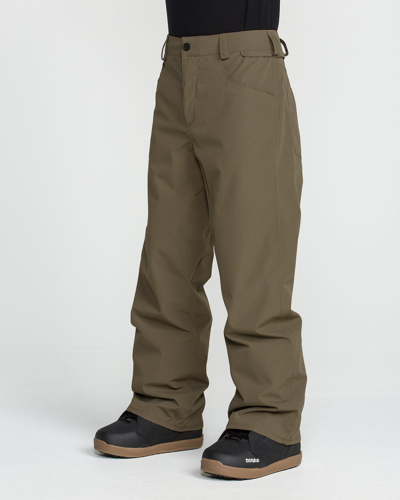 A person is shown wearing Volcom Men’s Nailer Pants in olive green, paired with black snow boots, standing against a plain white background. Only the lower half of the body is visible; these waterproof pants combine style and functionality for winter adventures.