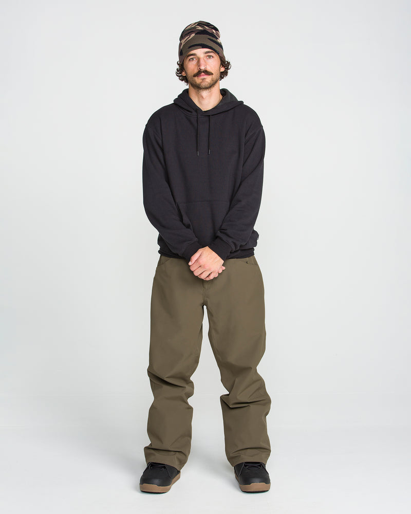 A person stands against a white background, wearing Volcom Men’s Nailer Pants in olive green with a baggy fit, paired with a black hoodie, black shoes, and a patterned beanie, hands clasped in front.