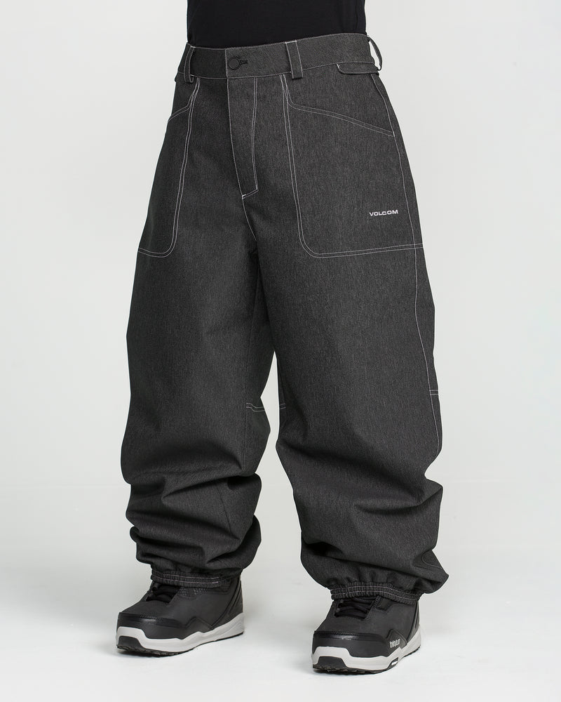 A person is wearing Volcom Men’s Lapper Pants in a loose-fitting, dark gray style with white stitching and gathered hems at the ankles. The pants feature the Volcom logo on the pocket and are paired with a black shirt and black and gray sneakers. The background is plain white.