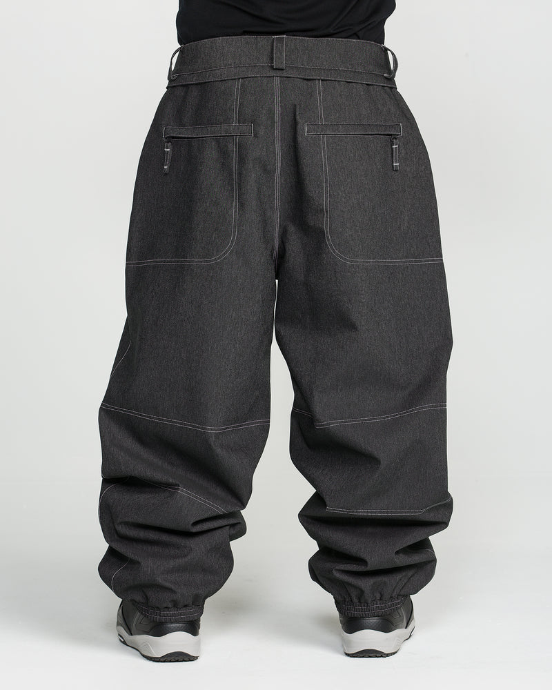 A person is wearing Volcom Men’s Lapper Pants in dark gray, featuring visible white stitching, large front pockets, and zippers, paired with gray sneakers and standing against a plain white background.