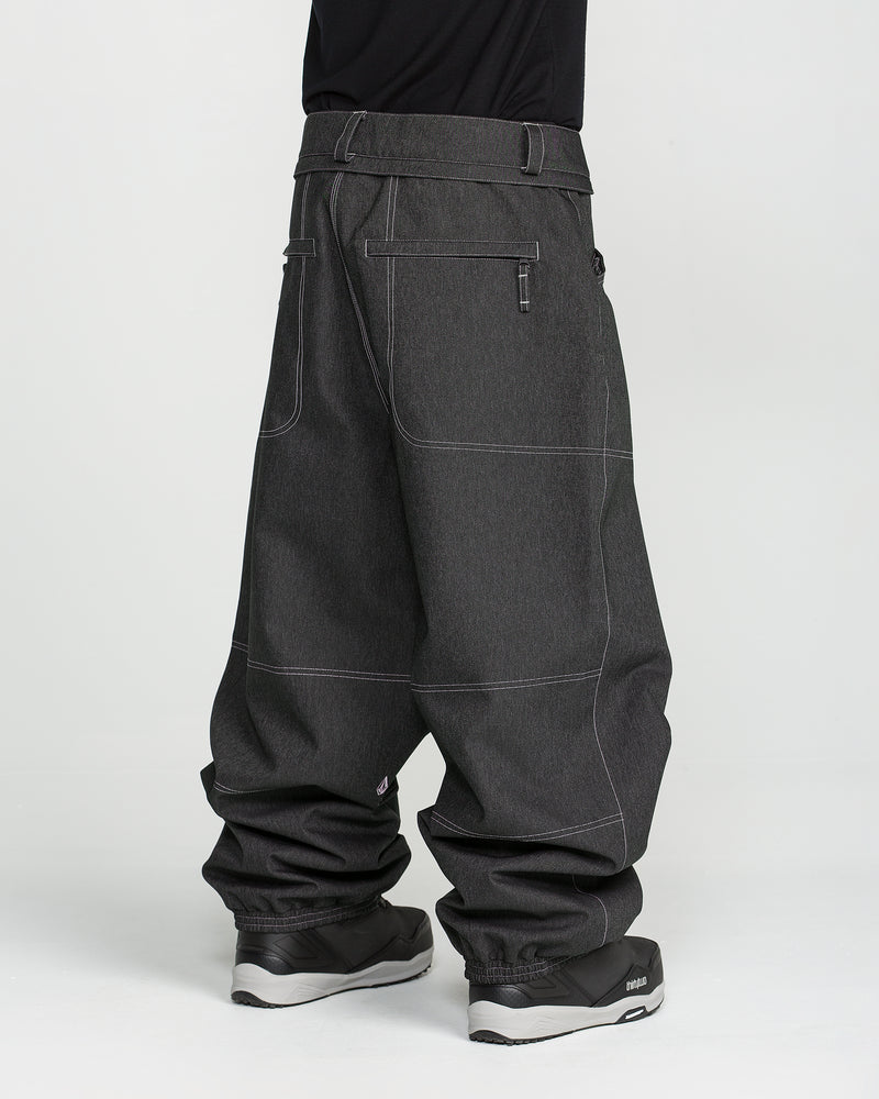 A person wearing Volcom Men’s Lapper Pants in dark gray, featuring white stitching, large pockets, and a loose, relaxed fit reminiscent of classic baggy snow pants, paired with black shoes and standing against a plain white background.