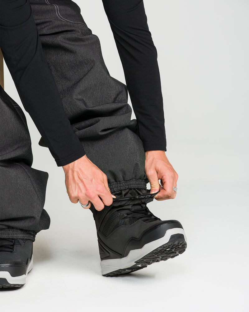 A person wearing Volcom Men’s Lapper Pants and black snow boots crouches down to adjust the cuff of their pant leg over the boot, set against a plain white background.