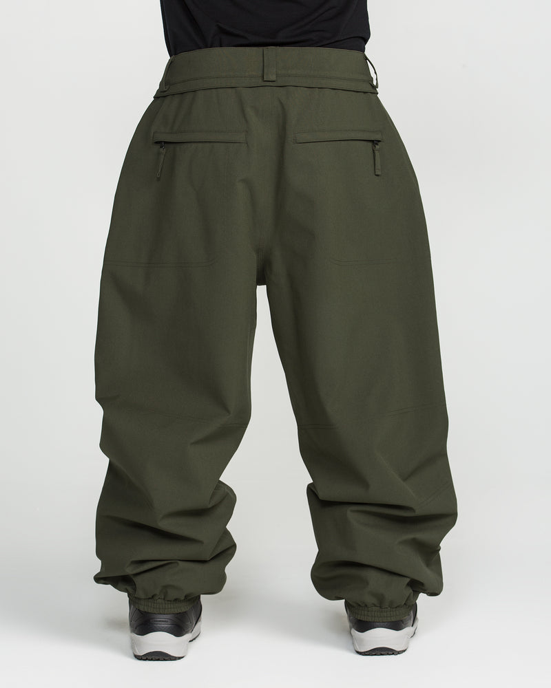 A person wearing Volcom Men’s Lapper Pants in olive-green, featuring zippered back pockets and elasticized cuffs, paired with sneakers, stands against a plain white background.