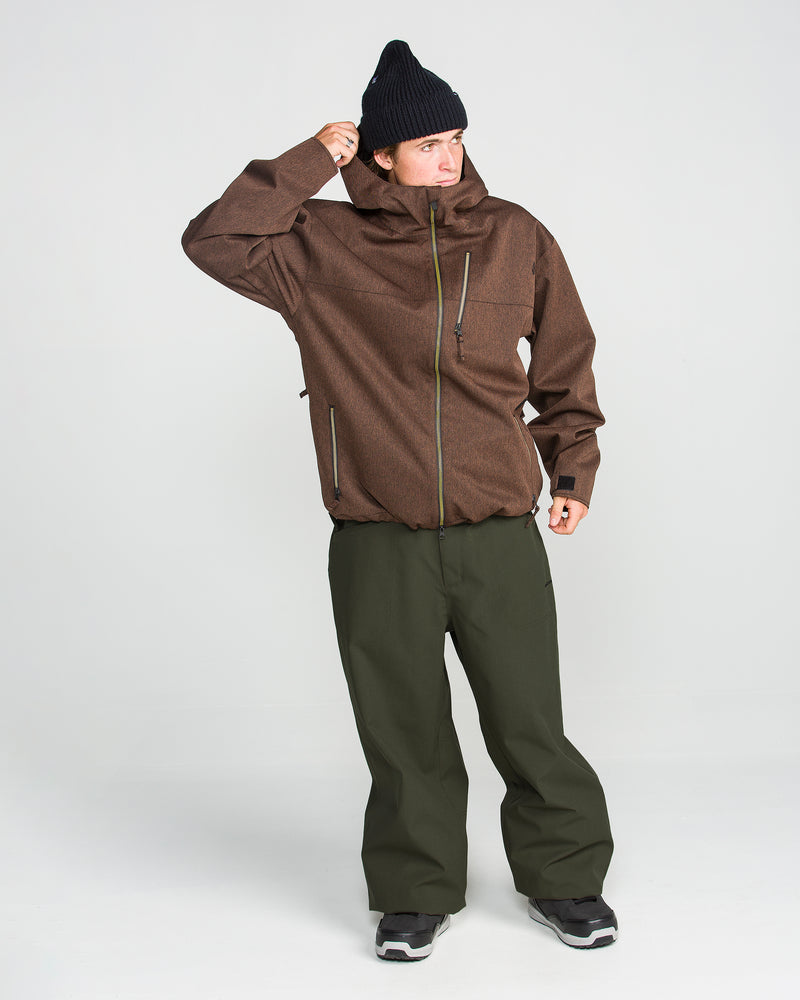 A person wearing a brown jacket, Volcom Men’s Lapper Pants in olive green, a black knit hat, and black shoes stands against a plain white background, adjusting their collar with one hand.