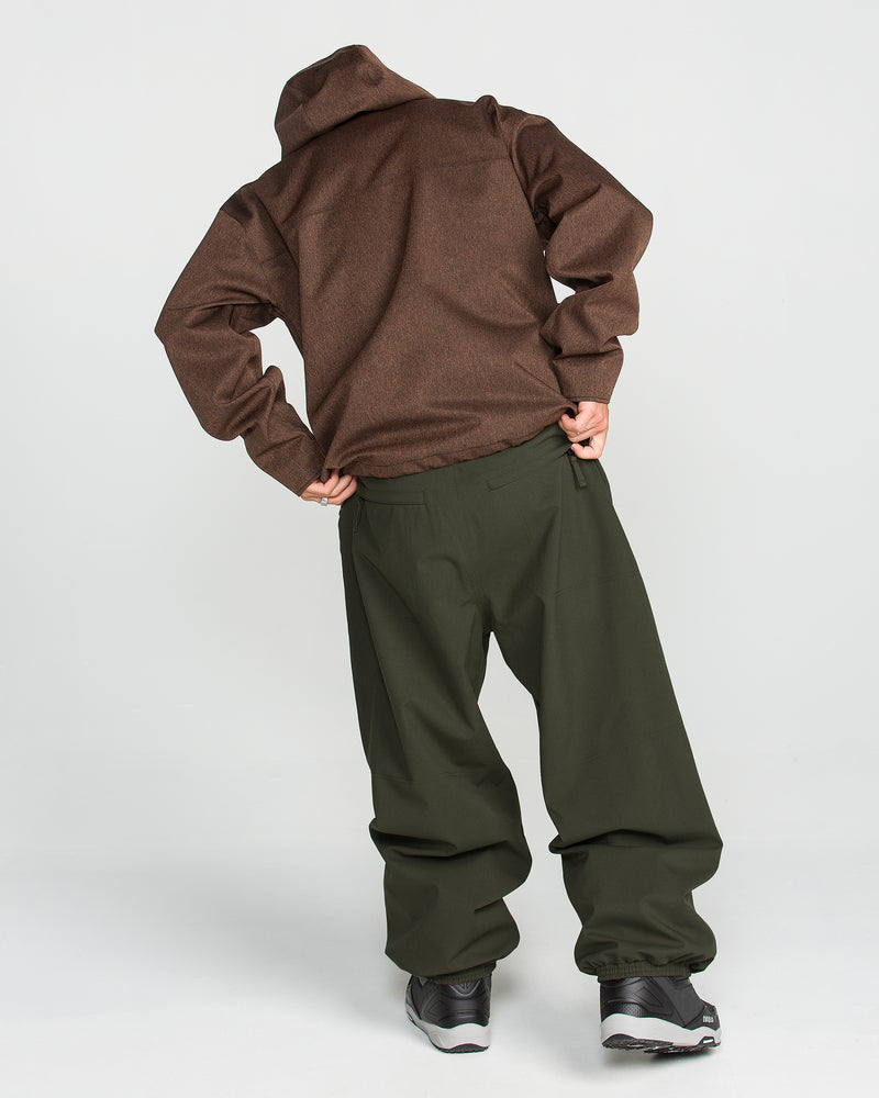 A person stands with their back to the camera, wearing a brown jacket, Volcom Men’s Lapper Pants in green, and black sneakers against a plain white background. Their hands rest on their hips as they lean slightly to one side.
