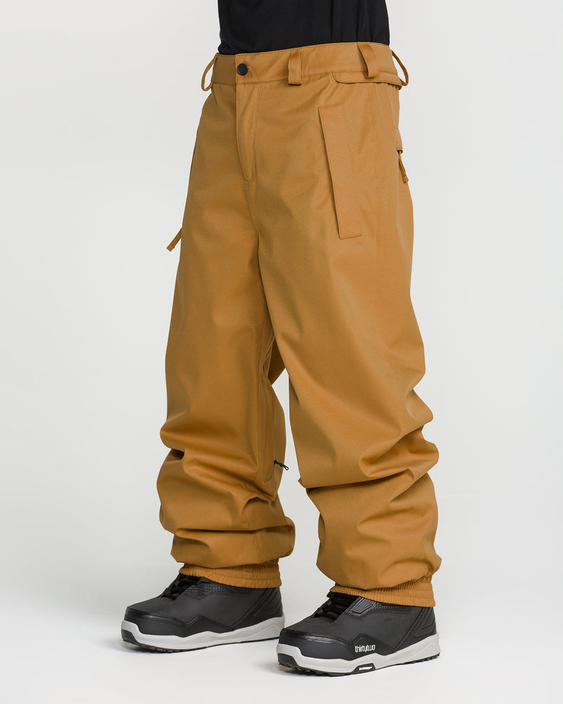 A person wearing loose-fitting tan Volcom Men’s Arthur 20K Pants, made from V-Science 2-Layer recycled twill, paired with black snow boots, stands against a plain white background with only the lower body visible.