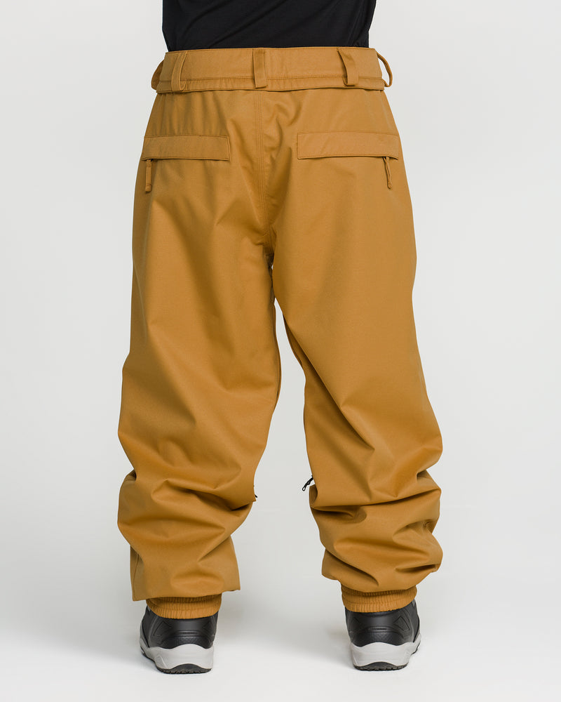 A person is shown from the waist down wearing Volcom Men’s Arthur 20K Pants—loose-fitting tan pants made from recycled twill featuring zipper pockets—paired with black shoes, standing against a plain white background.