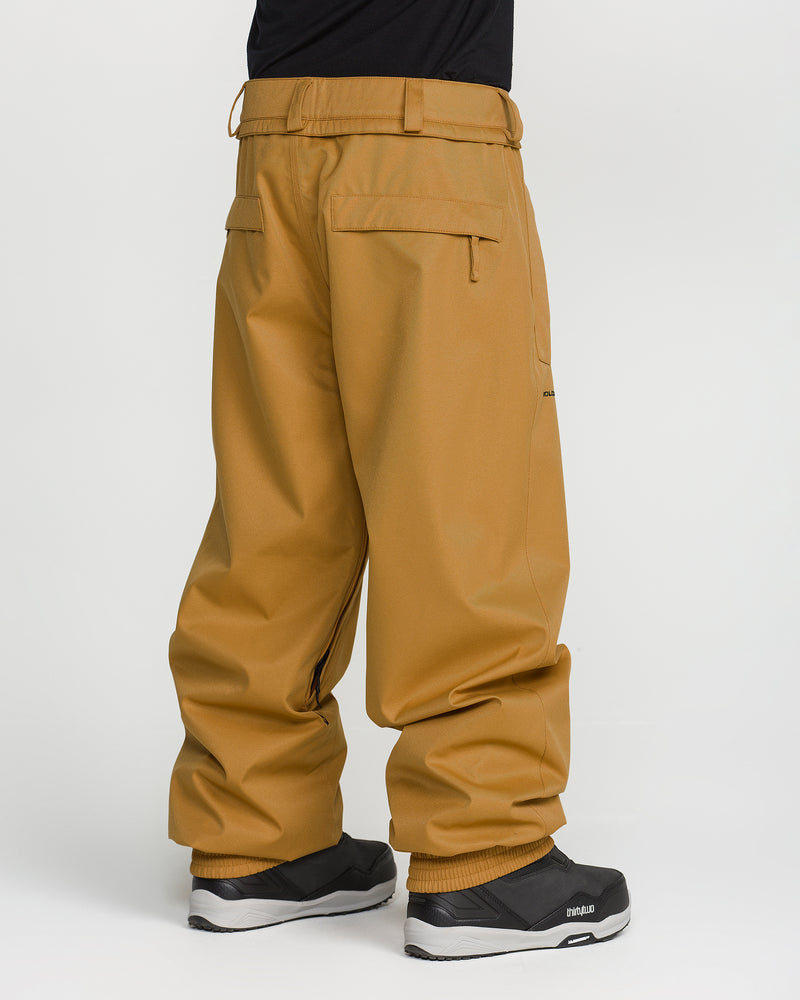 A person wearing loose-fitting tan Volcom Men’s Arthur 20K Pants, crafted from recycled twill with V-Science 2-Layer construction for comfort and durability, paired with black sneakers, is standing against a plain white background with their back to the camera.