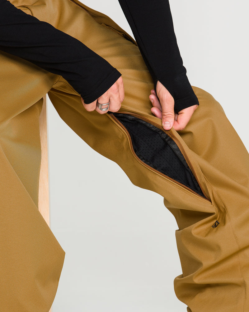 A person unzips a vent on the side of tan Volcom Men’s Arthur 20K Pants, revealing a black mesh lining inside. These pants feature recycled twill V-Science 2-Layer construction, inspired by the Arthur Longo 20K Pant. The person is wearing a long-sleeve black shirt.